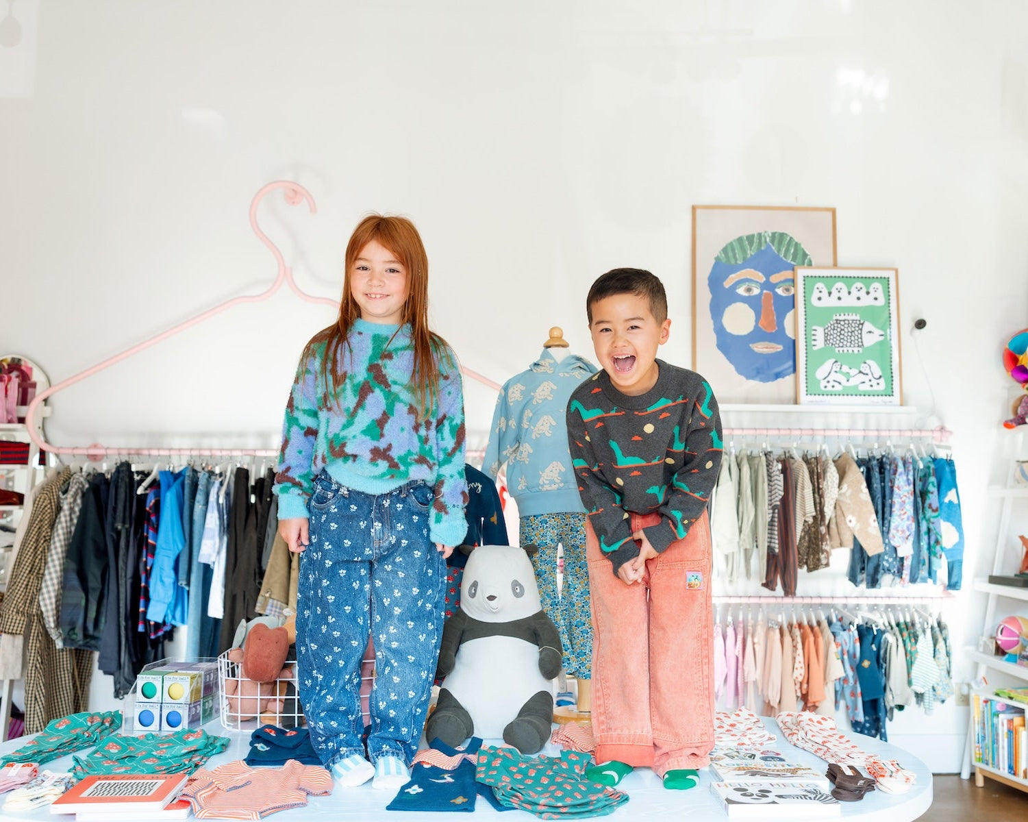 A young girl and a boy stand together in a bright, styled children's clothing boutique. The girl wears a teal and blue patterned long-sleeve top with matching blue star-print pants, while the boy wears a dark green patterned long-sleeve top with coral pink pants. at The Yo Store